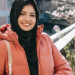 Cheerful ethnic female in hijab and jacket smiling and looking at camera on street