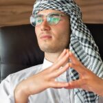 Middle Eastern man in professional attire with headscarf and glasses seated in a modern office environment.